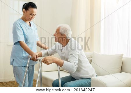 Healthcare facility. Positive nice delighted woman looking at her patient and helping him to walk while working as a caregiver