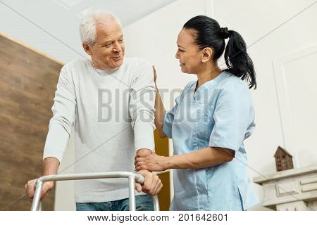 One more step. Cheerful positive friendly nurse standing near the elderly man and helping him to move around while being at work