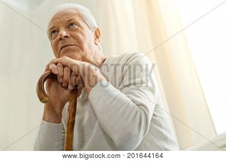 Old age. Thoughtful unhappy aged man holding a walking stick and leaning on it while thinking about his life