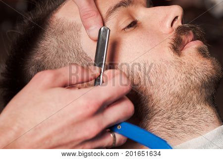 Hipster client visiting barber shop. The hands of young barber making the cut of beard