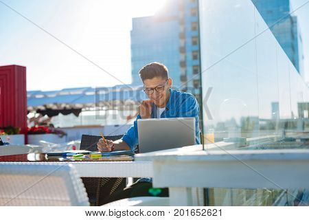 Positive thinking. Positive minded college guy smiling while looking at his home task and enjoying his time spent on a terrace of a local cafe.
