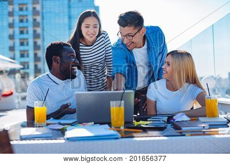 What is that. Curious young man looking at his friend and asking something while pointing at a screen of a laptop and working in a team on a terrace outdoors.