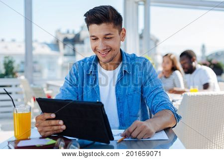 How is it possible. Happy self employed guy in casual smiling while looking at a screen of his tablet computer and reading news on the internet.
