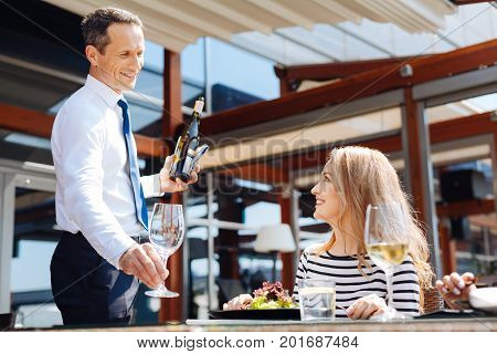 Restaurant service. Cheerful professional nice waiter putting a glass on the table and looking at his customer while preparing to pour wine