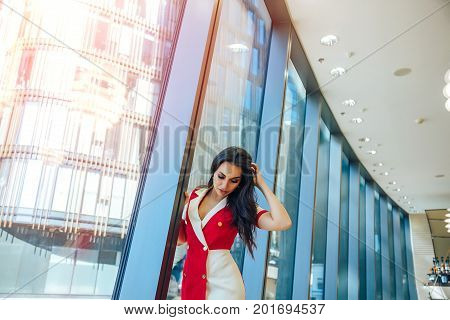 Young Beautiful Glamorous Brunette Girl In A Red Dress Posing Near Glass Showcase In Business Center