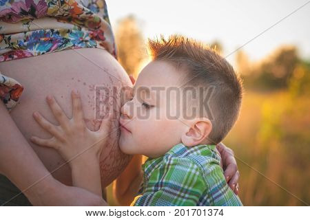 Cute boy kissing and listening his mothers pregnant mehandi pattern ornament belly on sunset nature landscape