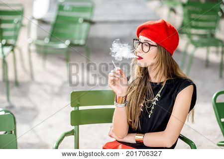 Young stylish woman in red cap smoking a cigarette sitting outdoors on the green chairs in Paris
