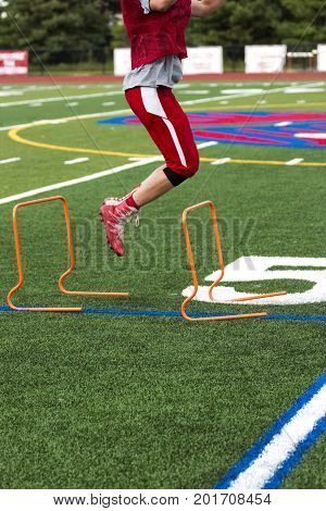 A wide receiver performing plyometrics by jumping over orange hurdles on a green turf field at football practice