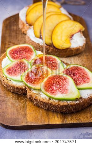 Pouring honey on toasts with cream cheese topped with fresh ripe figs and peaches. Wholegrain rye bran bread on wood cutting board grey concrete background.