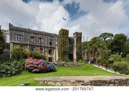 Clifden Ireland - August 4 2017: The historic gray and brown stone castle of the town is now an upscale hotel. Ivy on the walls in flower setting on hill with forest against blue sky with white clouds.