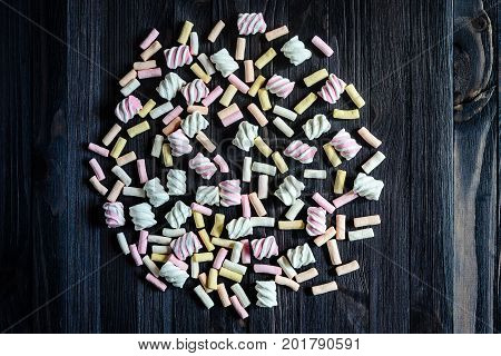 Laid out in the shape of a circle multicolored marshmallows on black wooden table top view. Background of multi-colored and different in shape and size of marshmallows. Sweets background