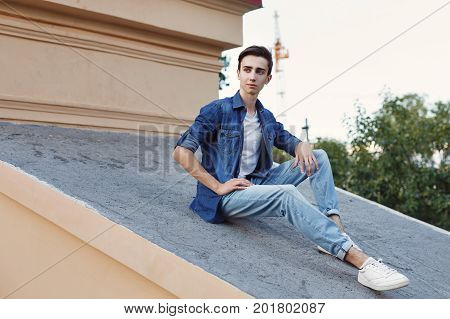 A young guy is sitting on the roof. He is wearing a jacket and jeans. Hipster city fashion.