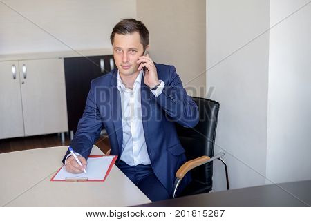 serious business man in office making cell phone call and taking notes.