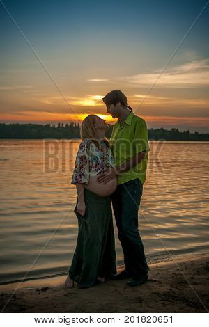 A pregnant beautiful woman with her husband on the river bank beach smiling and touching her mehandi ornament belly with love and care. Happy couple, family relax on sunset