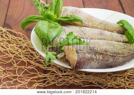 Fragment of a white dish with carcasses of the notothenia fish without of a heads and tails and with peeled scales basil and parsley twigs on a dark wooden surface with fishing net closeup