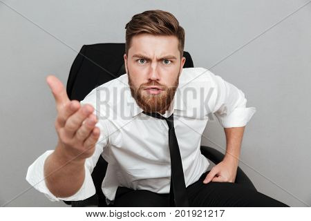 Close up portrait of a young unsatisfied man in white shirt staring at camera and pointing at camera while sitting in chair isolated over gray background
