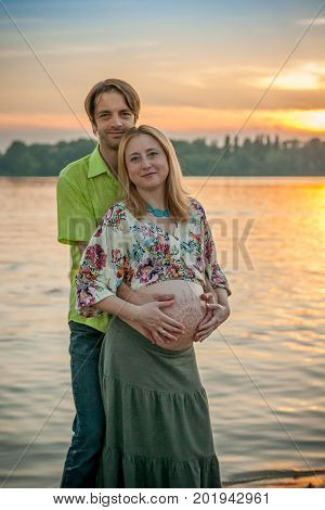 A pregnant beautiful woman with her husband on the river bank beach smiling and touching her mehandi ornament belly with love and care. Happy couple, family relax on sunset