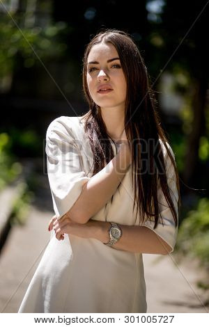 Young Brunette Woman In The Park Touching Her Neck With Her Right Hand. She Is Wearing A White Dress
