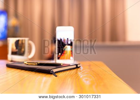 Notebook And Pen With Cell Phone On Wooden Table In Hotel Room.