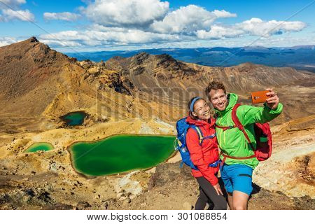 New Zealand Tongariro Alpine Crossing Hiking tourists couple selfie at Emerald Lakes. Happy backpackers tramping taking phone photo of themselves at volcanic mountains. Tramping track of New Zealand.