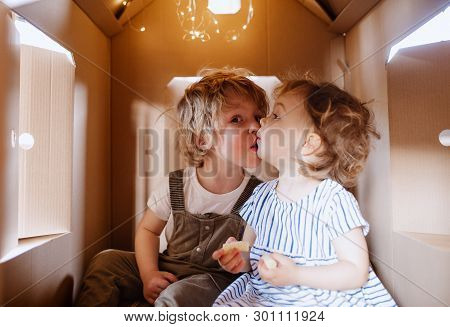 Two Toddler Children Playing Indoors In Cardboard House At Home, Kissing.