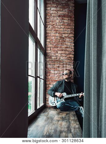 Man Singer Sitting On A Window Sill In A Headphones With A Guitar Recording A Track In A Home Studio