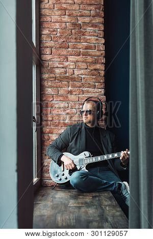 Man Singer Sitting On A Window Sill In A Headphones With A Guitar Recording A Track In A Home Studio