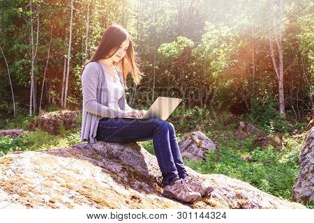 Young Beautiful Girl Is Working On A Laptop While Sitting On A Big Rock In The Forest. Freelance Con