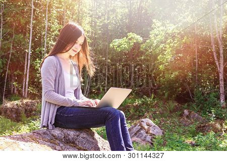 Young Beautiful Girl Is Working On A Laptop While Sitting On A Big Rock In The Forest. Freelance Con