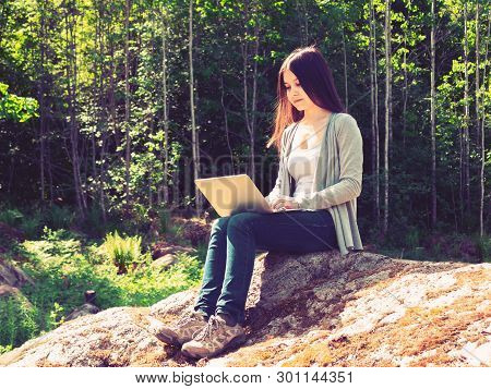 Young Beautiful Girl Is Working On A Laptop While Sitting On A Big Rock In The Forest. Freelance Con