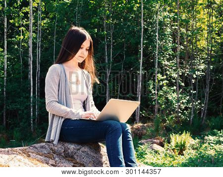 Young Beautiful Girl Is Working On A Laptop While Sitting On A Big Rock In The Forest. Freelance Con