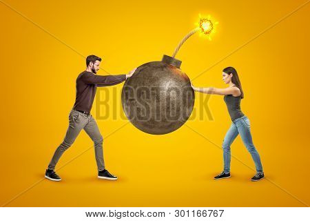 Young Man And Woman In Casual Clothes Holding Big Ball Bomb With Fuse On Yellow Background