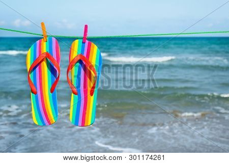 closeup of a pairs of colorful flip-flops, patterned with the colors of the rainbow flag, hanging on a clothes line on the beach, with the sea in the background and a blank space on the right