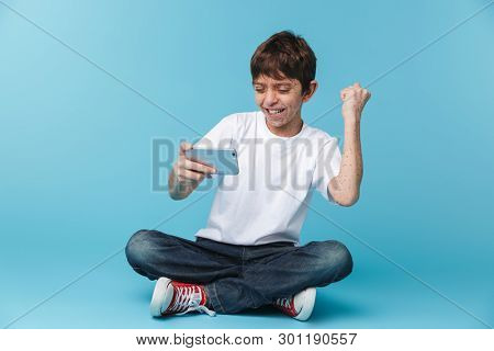 Image of pleased brunette boy 10-12y with freckles wearing white casual t-shirt holding and using smartphone while sitting on floor isolated over blue background