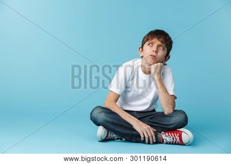 Image of european youngster boy 10-12y with freckles wearing white casual t-shirt looking upward while sitting on floor isolated over blue background