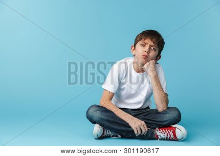 Image of caucasian brunette boy 10-12y with freckles wearing white casual t-shirt looking at camera while sitting on floor isolated over blue background