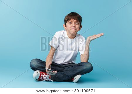 Image of teenage brunette boy 10-12y with freckles wearing white casual t-shirt holding and using smartphone while sitting on floor isolated over blue background