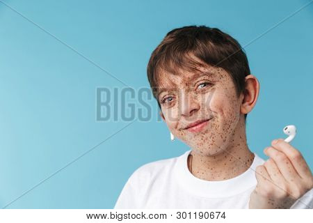 Image of beautiful caucasian boy 10-12y with freckles wearing white casual t-shirt and earpods smiling at camera isolated over blue background
