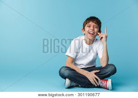 Image of young brunette boy 10-12y with freckles wearing white casual t-shirt showing finger upward while sitting on floor isolated over blue background