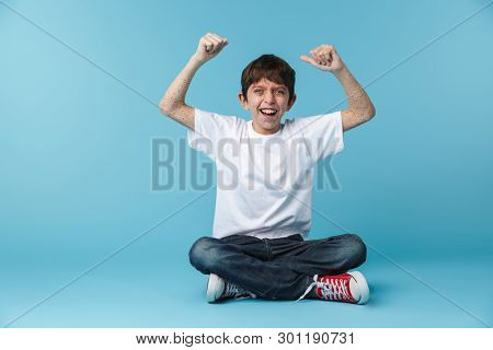 Image of happy brunette boy 10-12y with freckles wearing white casual t-shirt smiling at camera while sitting on floor isolated over blue background