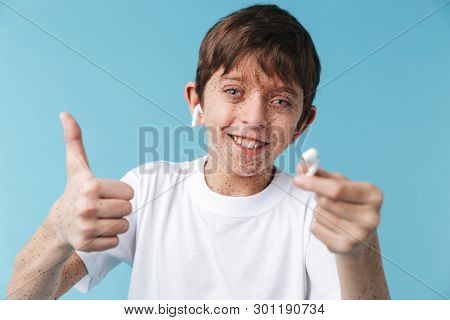 Image of charismatic caucasian boy 10-12y with freckles wearing white casual t-shirt and earpods smiling at camera isolated over blue background
