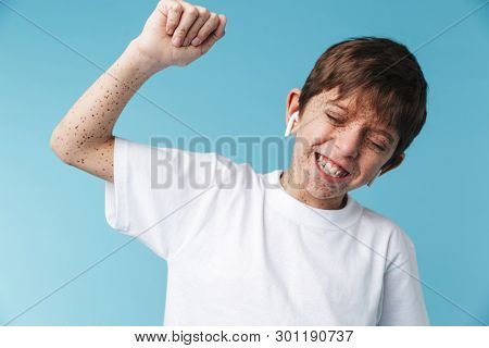 Image of joyful caucasian boy 10-12y with freckles wearing white casual t-shirt and earpods smiling at camera isolated over blue background