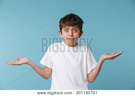 Portrait closeup of puzzled beautiful boy 10-12y with freckles wearing white casual t-shirt looking at camera isolated over blue background