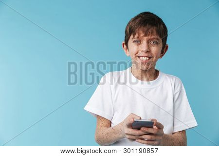 Image of cheery caucasian boy 10-12y with freckles wearing white casual t-shirt smiling and holding smartphone isolated over blue background