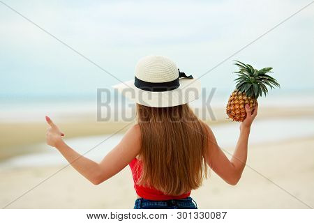 A Young Girl In A Light Hat Stands With Her Back And Holds A Pineapple In One Hand.