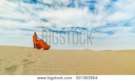 Girl Walks On Desert In Dress Fluttering In Wind. Beautiful Woman Is Walking, Staing On Sand Or Dune