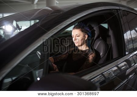 Woman Is Testing New Auto In Car Showroom. Beautiful Blue-haired Girl In Black Dress Is Driving Luxu