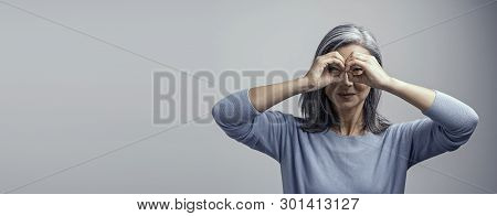 Beautiful Grey-haired Woman Squints Her Eyes While Looking Through Her Hands As If They Are Glasses.