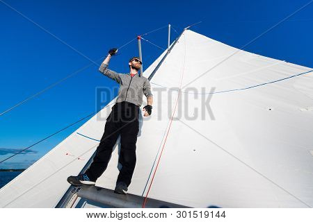 Yacht Captain With A Beard Stands On Sail Boom On A Sailing Yacht, Holding The Rope In His Hand And 