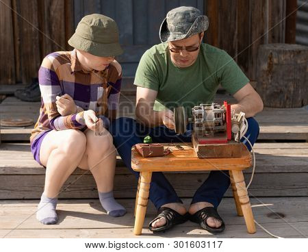The Caucasian Man And The Woman Are Sitting On Wooden Porch Of Their House And Working With The Port